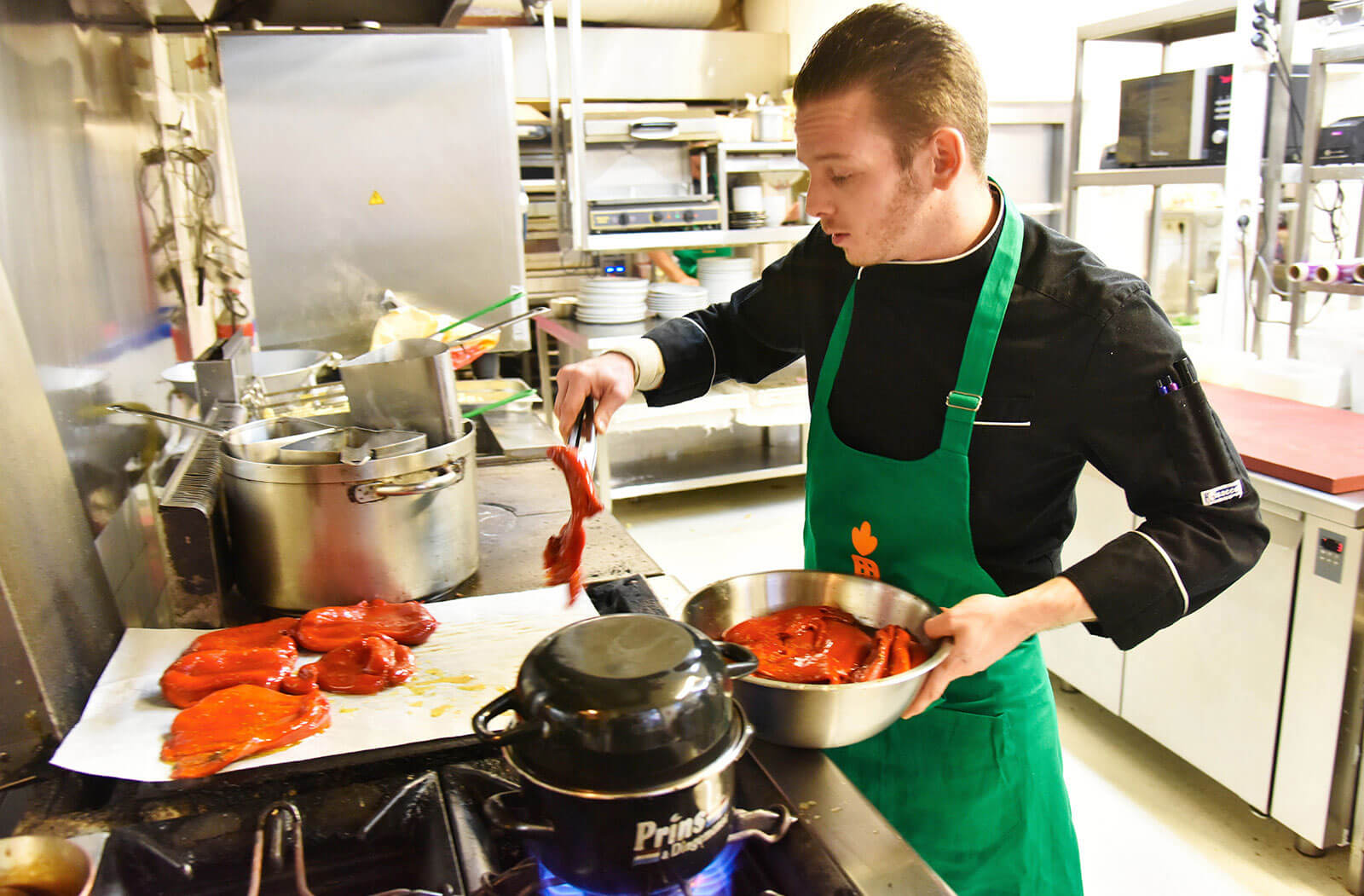 Dans une cuisine de collectivité, un homme met des morceaux de viande dans un plat. Il porte un tablier vert avec une carotte orange.