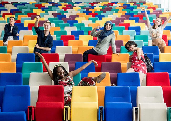 Six personnes qui posent de manière humoristique dans un auditorium, assis sur des fauteuils multicolores.
